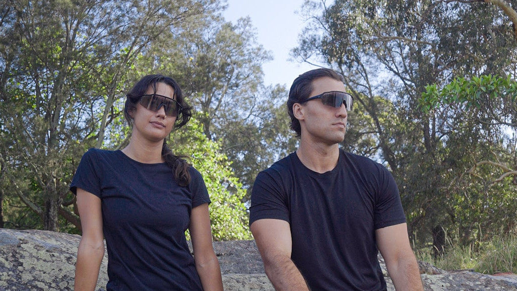Two people sitting on a log in a forested area with sunglasses
